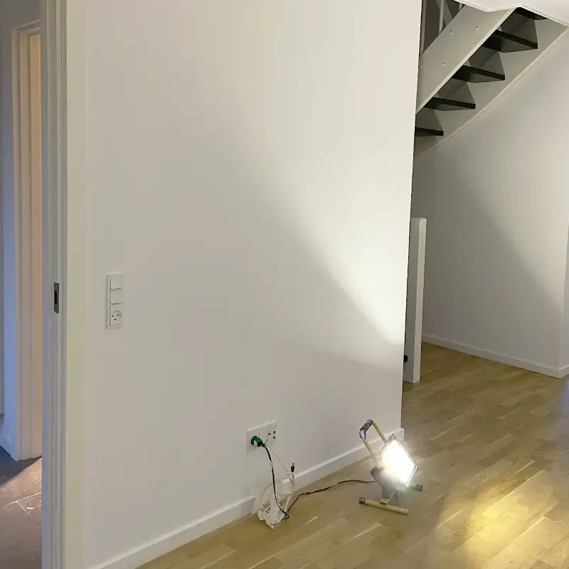 Nymalet hvid væg i entré med trappe og lyst trægulv oplyst af arbejdslampe